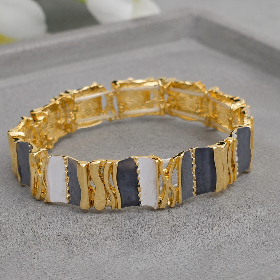 A gold-plated stretch bracelet with alternating rectangular gold and dark gray inlay segments, displayed on a gray jewelry stand with blurred white flowers in the background.