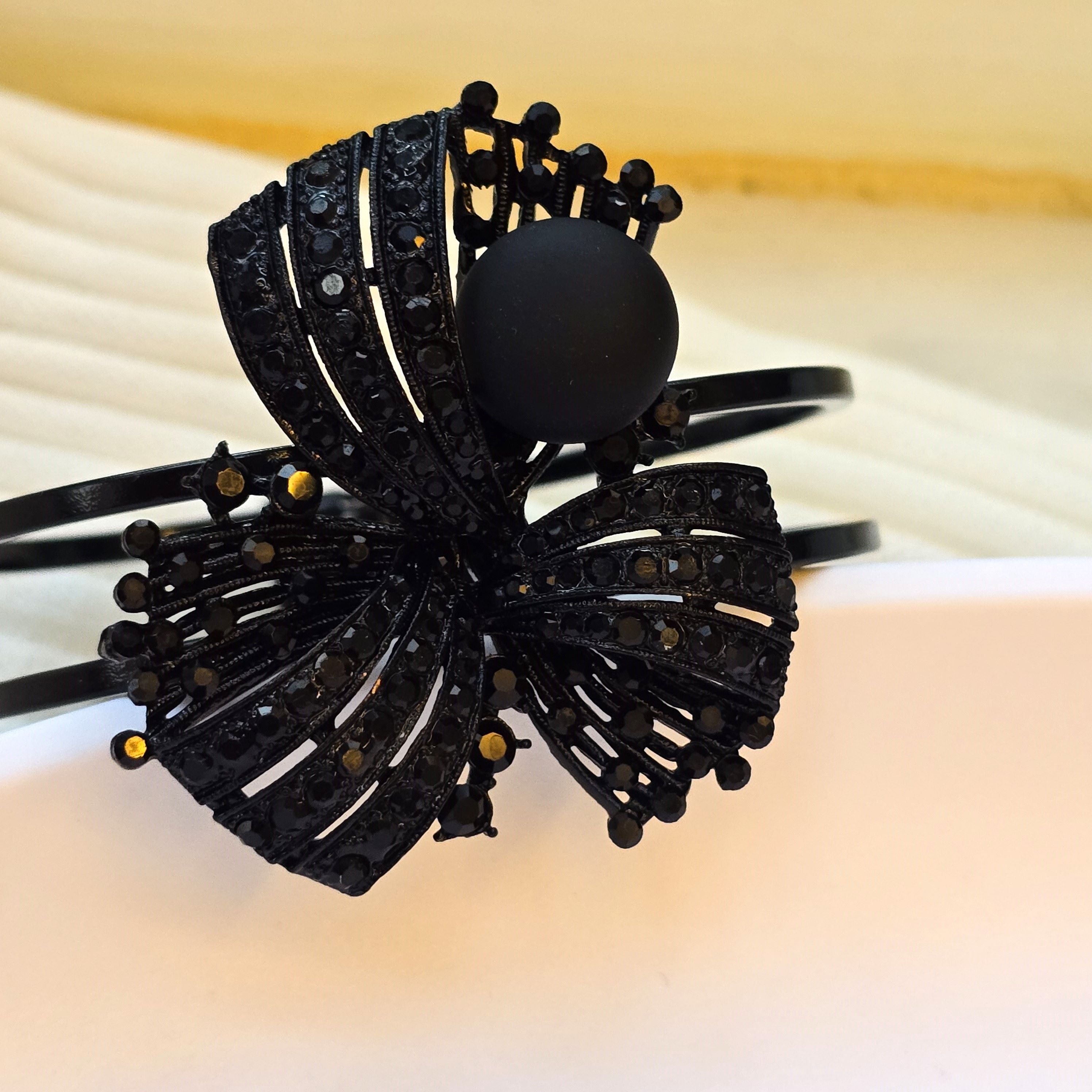 A black metal wrap bracelet featuring a large, ornate bow design embellished with black rhinestones and a central matte black oval stone.