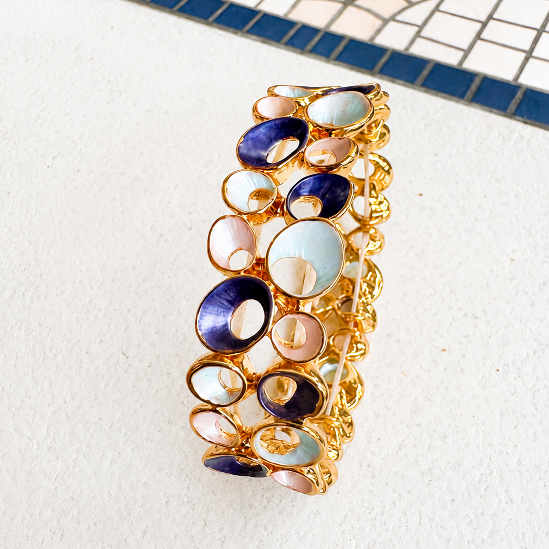 A gold-plated bangle bracelet featuring multiple oval and circular links filled with colorful enamel in shades of blue, green, resting on a white textured surface with a blue and white mosaic tile pattern in the background.