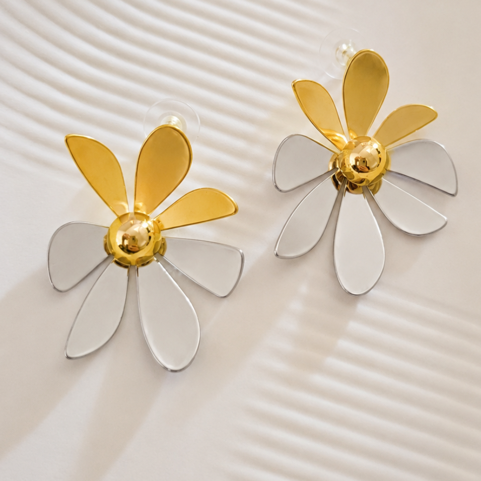 A pair of gold and white flower-shaped stud earrings displayed on a textured, light beige surface with concentric wave patterns.
